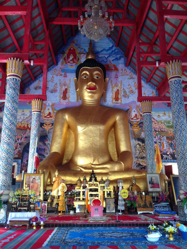 The Buddha sits cross-legged in a massive sculpture. He has bright gold skin, red lips, elongated earlobes, and a gold topknot. Framed images of monks and flower offerings have been left in front of him.