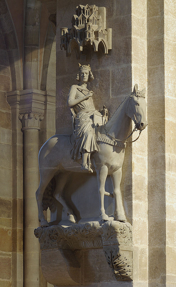 A leafy, carved pedestal on a stone pillar holds a pale stone sculpture of a horse with a regal rider. The rider wears a crown and draping robes and cloak. A sculpture of castle in miniature is affixed to the wall above the group.