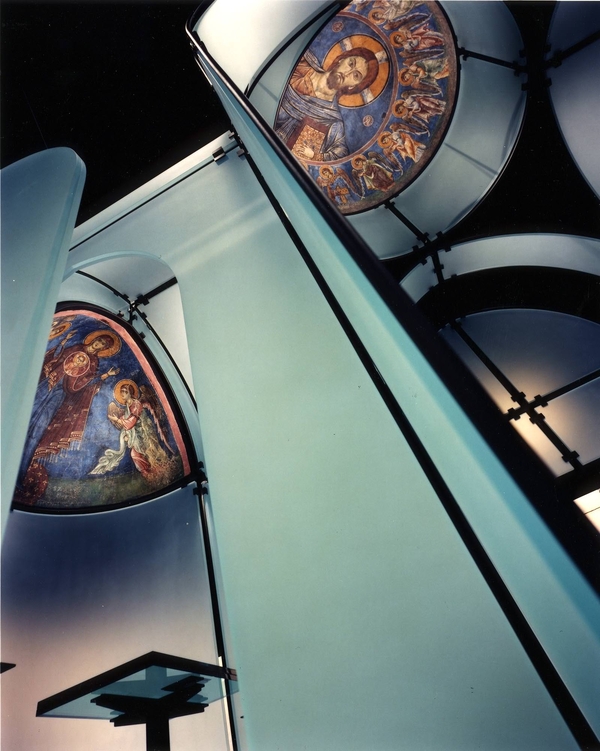 A photograph taken from the base of a floating glass wall captures the painted blue Byzantine frescos of Christ pantocrator and angels hung in the dome and apse of the glass architectural structure.