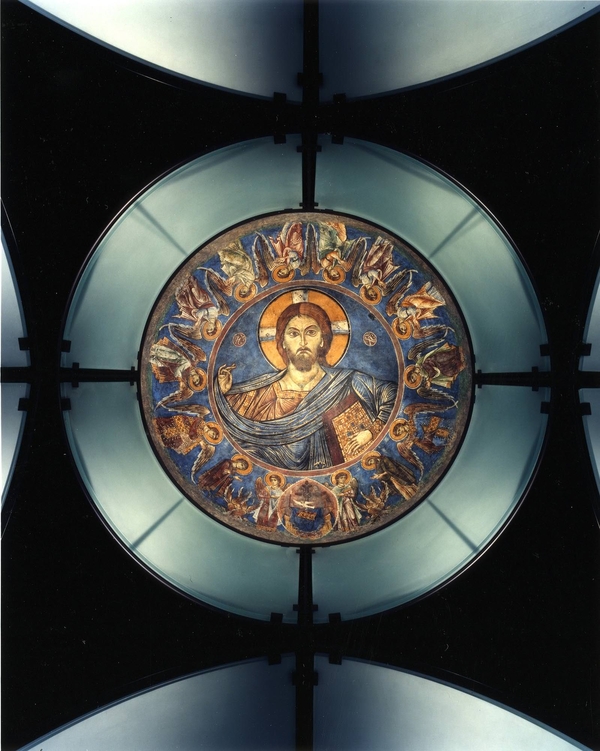 A Byzantine blue fresco installed in a glass and steel architectural dome depicts a light-skinned Christ from the bust up and encircled by a row of winged angels. Christ holds up his right hand and grasps a book in the left. He looks out sternly. 