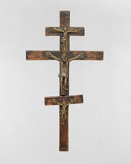 A wooden object consists of three crucifixes stacked vertically. A cast metal, Christ figure is affixed to each crucifix. Their hands are outstretched, their waists covered with loin cloths, and their ribs visible.