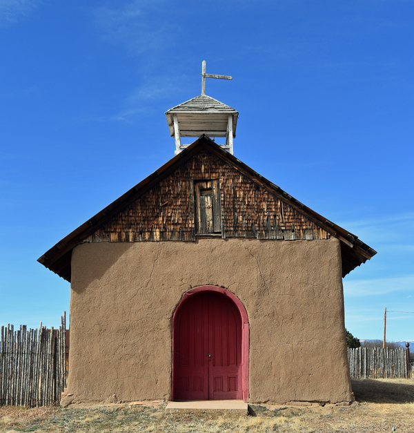 On an otherwise muted adobe church, a bright red door at the entrance stands out. The building has a gable roof with wooden shingles and a very short, dilapidated gabled bell tower on top. It holds up the remains of a cross.