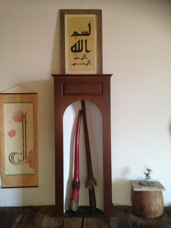 A collection of wall-hangings and framed prints of Arabic text are arranged among other wooden art objects and furniture pieces.