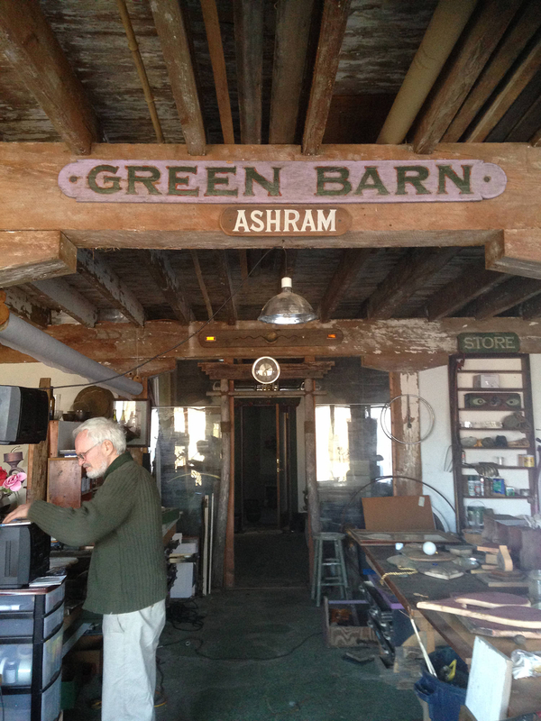 An older, light-skinned man rifles through objects in a barn work space. Two signs affixed to a projecting ceiling beam read, "GREEN BARN" and "ASHRAM."