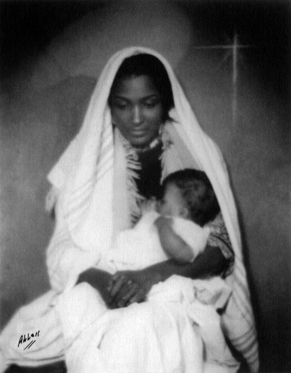 A black and white photo depicts a dark-skinned woman in a white veil cradling a swaddled baby close to her chest. She fold her hands in her lap and casts her eyes down. A nativity star is visible in the righthand corner.