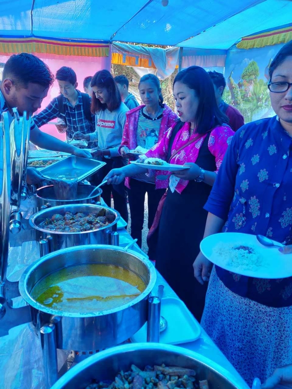 A line of South Asian men and women move along a buffet-style table set up with vats of food, and they spoon the meal onto their plates.