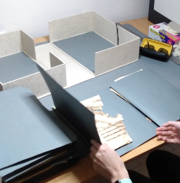 Document boxes on a table, hands at bottom of image handle a box containing a manuscript fragment
