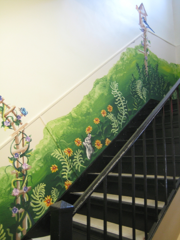 The wall beside a staircase is painted with an idyllic garden mural that includes trellised flowers, green grass, a bird feeder, and a bunny among the flowers.