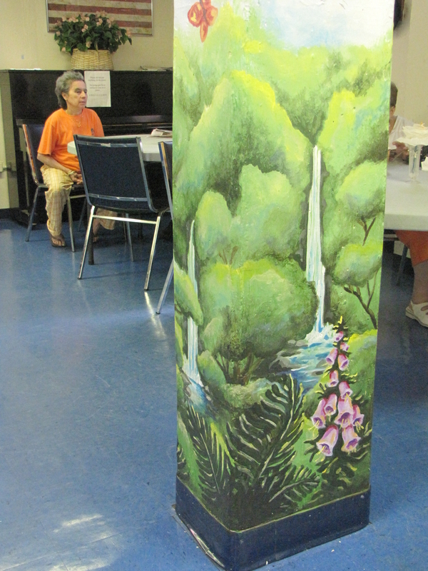 A mural of trees, flowers, and a flowing waterfall wraps around square post. The post stands in a simple interior with plastic tables and chairs.