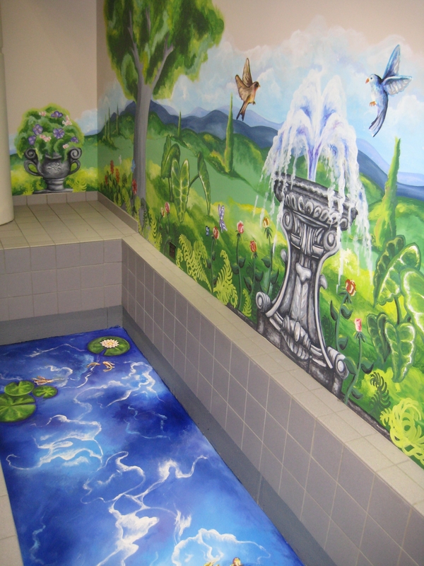 A mural in a tiled interior depicts a birdbath fountain that bubbles over with water. The mural includes trees, flowers, and fluttering birds. A second mural of blue water and lilypads is painted in a recess in the ground before the first. 