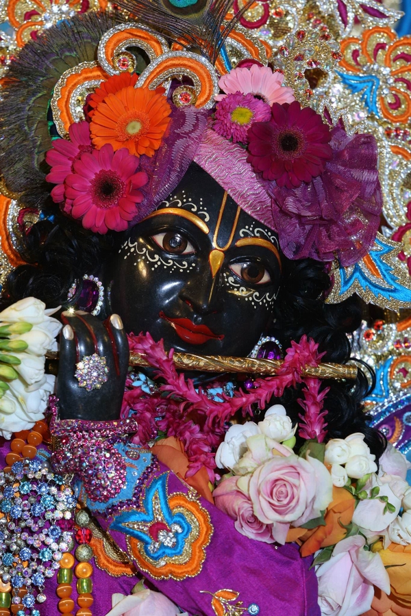In this close-up photograph, a black-colored figure raises a garlanded flute to their ruby red lips. They have big brown eyes, golden facial markings about their eyes and on their nose bridge, and a floral headdress.