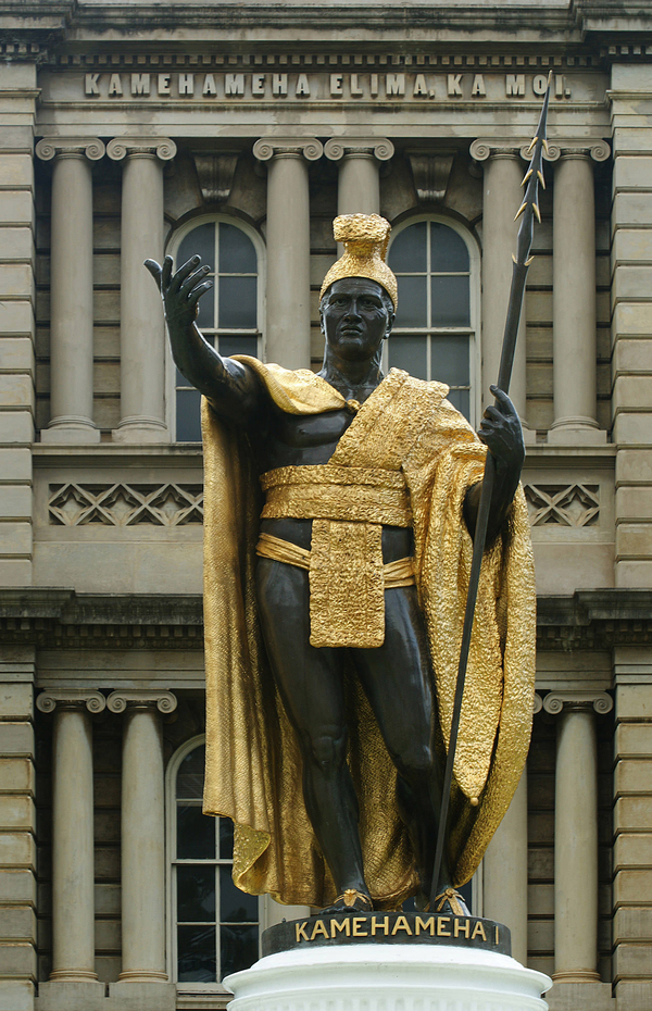 A heroic, oversize statue of a male figure has a dark patina and muscular body. He wears a golden gilt helmet, sash, and cloak. The figure adopts a contrapposto stance and raises his left arm while holding a spear in the right.