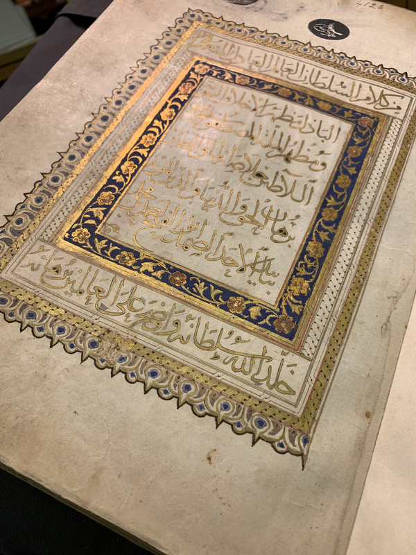 An illuminated Islamic manuscript is open to its cover page with gold Arabic calligraphy. The text is framed by multiple blue and gold borders of flowers and interlace.
