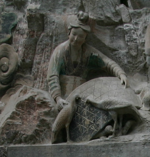 A woman sculpted of gray stone emerges from a rocky outcropping. She reaches her arms out over a sculpted basket with two thin birds in front of it and small chicks underneath it. 