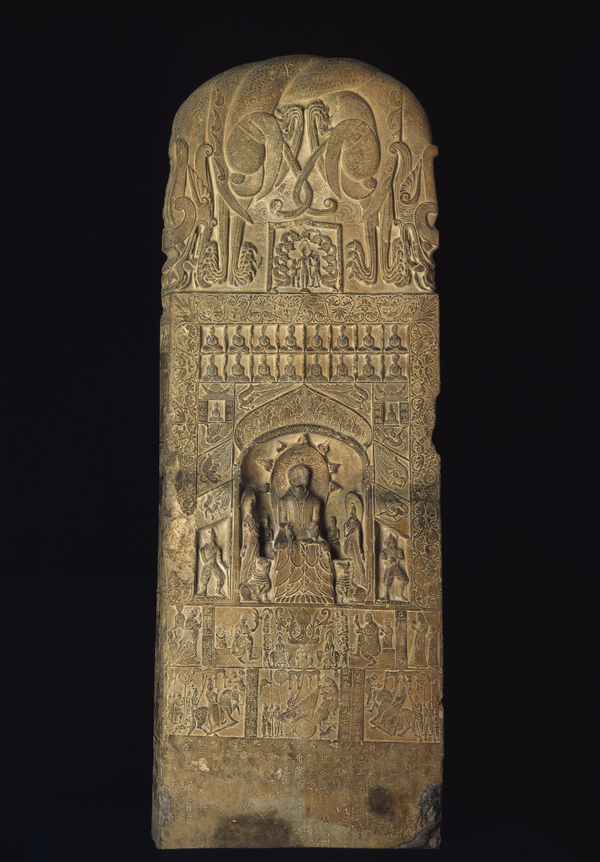 A pale stone slab is covered with low relief carvings. A central niche holds an ethroned figure carved in higher relief among his attendants. A variety of figures and processional scenes surround him as well as abstract vining and floral motifs.