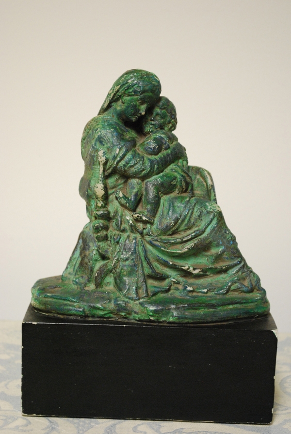 A plaster figurine depicts Mary cradling the Christ child close. Shown in profile, Mary wears a draping robe that renders her into a throne for her chubby son. The entire group is weathered and greenish.