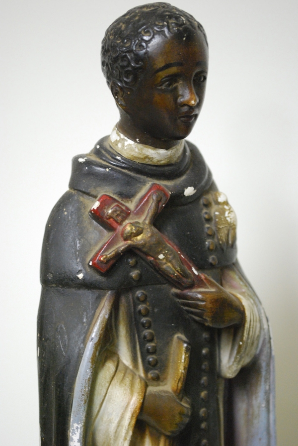 A close-up of a plaster statue shows a dark-skinned figure grasping a red and gold crucifix close to his chest. He wears brown and white robes and has close-cropped brown hair.