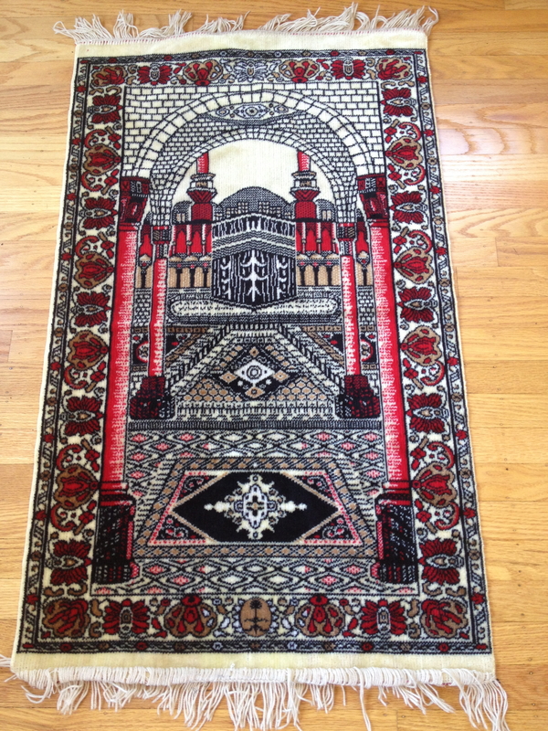 A prayer rug depicts the black, square Kaaba woven in an ornate red, white, and black architectural space. The setting includes columns, arcades, and patterned rugs.