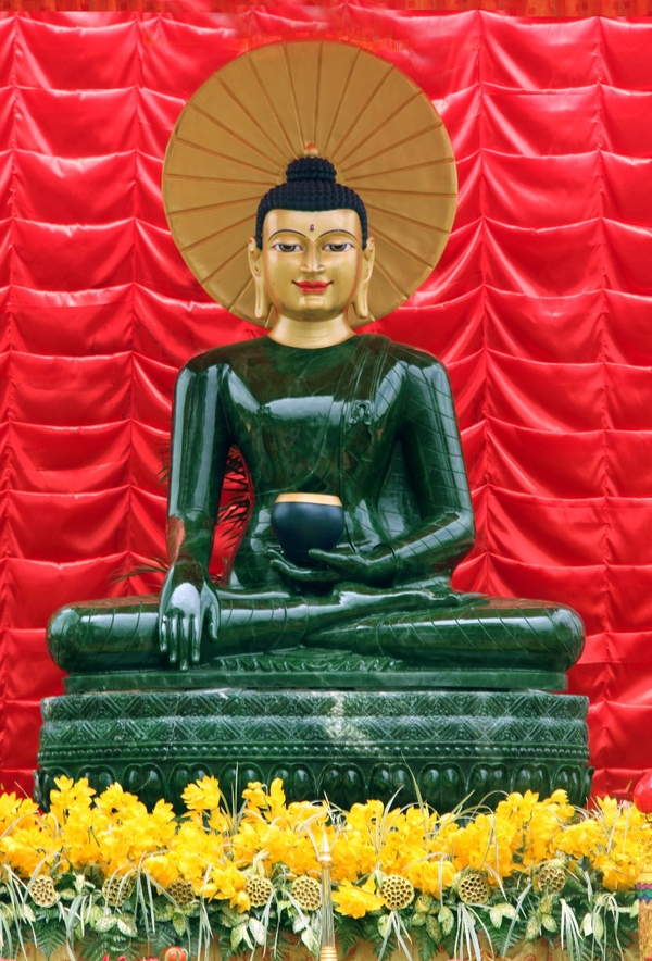 A large, shiny jade figure sits cross-legged on a carved platform. The head is gold with large eyes, long earlobes, a black topknot, and serene expression. It holds a small black object. The figure sits against a red background and among yellow flowers.
