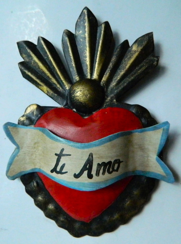 A small red metal heart is fashioned with a white and blue banner unfurled across it. The banner is inscribed with "te Amo" in black, handwritten script. Abstract, metal flames emerge from the top of the heart. 