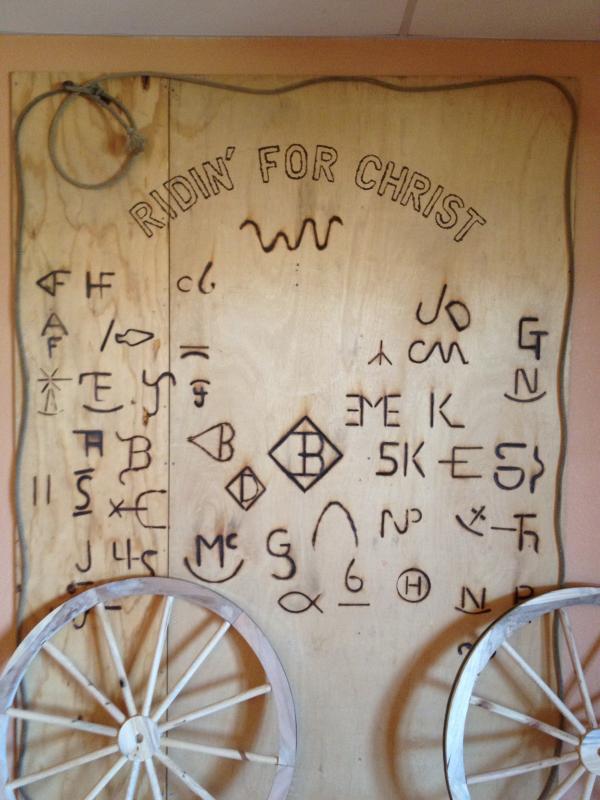 A slab of wood is branded with a variety of symbols and text reading, "RIDIN' FOR CHRIST." Two wagon wheels are affixed to the bottom of the wooden slab.