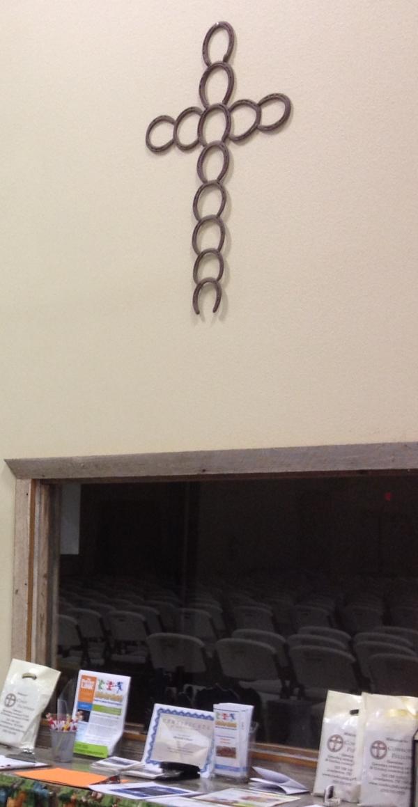 Horseshoes have been linked end to end in a cruciform shape. It hangs on a plain wall above a table arrayed with brochures.