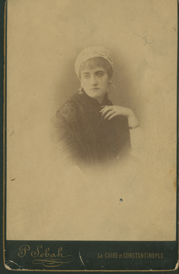 A woman with a light-colored headscarf and dangling earrings looks out to the left of a sepia portrait photo. She raises her right hand to her chest in an elegant gesture. There is a bangle on her arm.