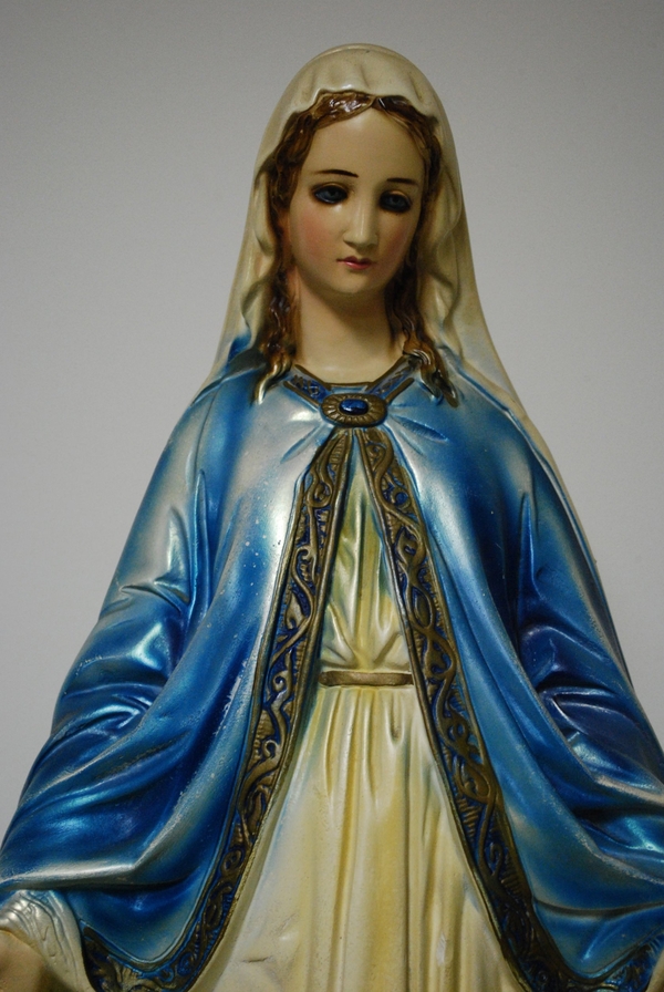A light-skinned chalkware Marian figure wears a white veil and a blue, spray-painted cloak. The cloak has a golden, decorative border and jeweled clasp. The woman stretches out her hands and casts her eyes down.