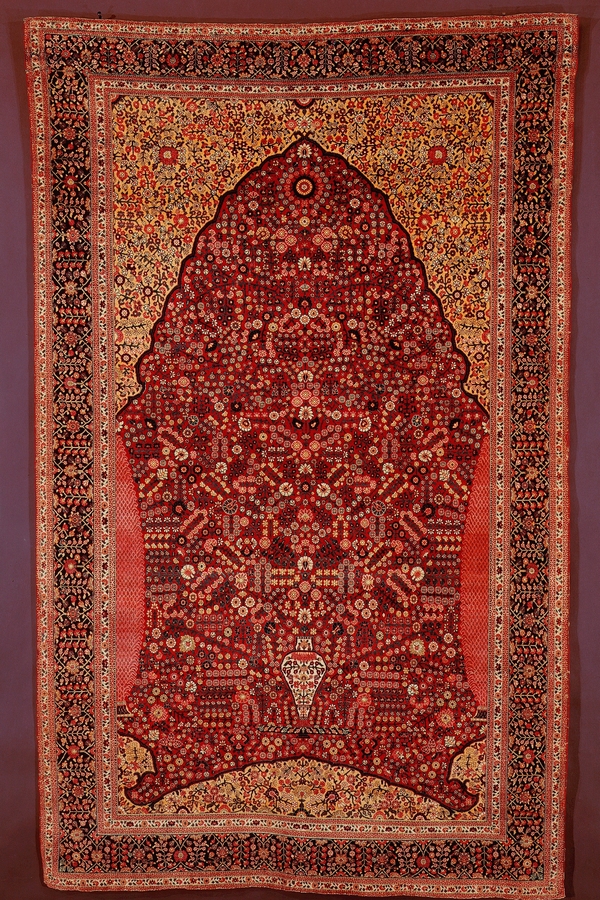 An ornate red rug is woven all over with small, geometric floral motifs. A red mass of flowers emerges from a small white vase represented within a gateway shape.