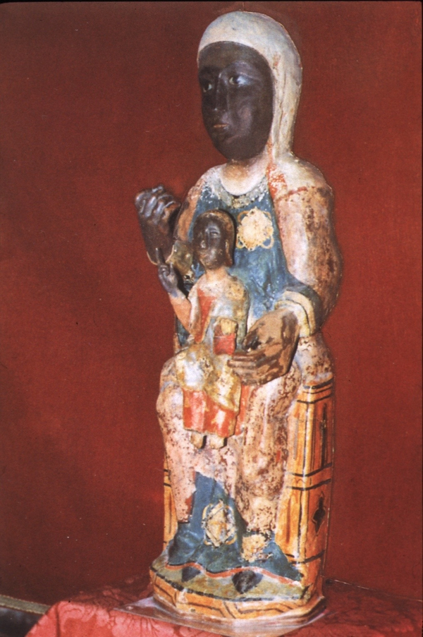 A sculpture of a dark-skinned woman depicts Mary enthroned with a small, dark-skinned child on her lap. Both raise their right hands in blessing. Much of their clothes and the throne are faded and chipped from time.