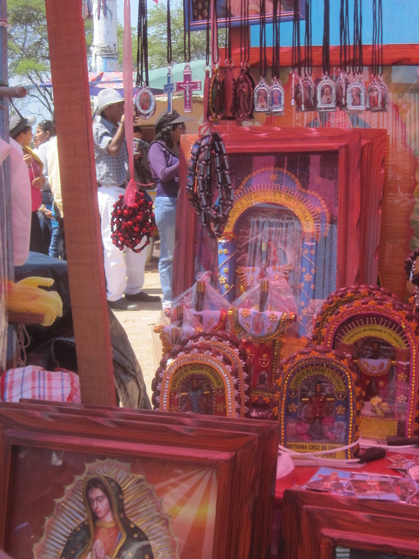 Souvenirs assembled on a table and hanging from a stall include crucifix pendants, paintings of Our Lady of Guadalupe, and other colorful icons covered with decorative beads.