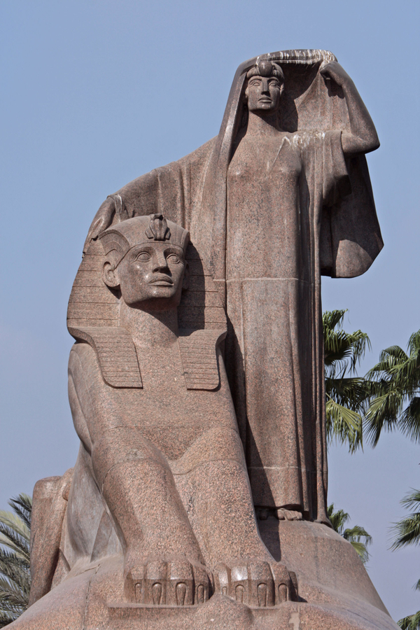 A columnar woman in long robes wraps her hand around a sphinx in a large sculpure. The sphinx has oversized paws, a masculine human face, and a pharaonic headdress. 