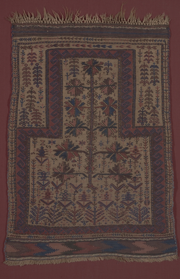 A muted, ruddy colored rug depicts an abstract tree. The tree is framed by a patterned border and surrounded by abstract, vegetal motifs.
