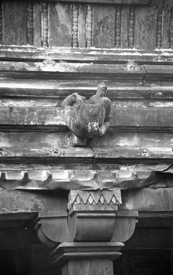 A grayscale photo of the entablature above an ornate stone pilaster includes a protruding female torso. She places a hand on her necklace-draped chest.