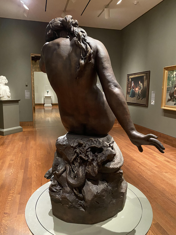 A naturalistic nude bronze of Eve is viewed from behind. She sits upon a rock around which a scaly snake curls. The woman cowers with her arm stretched back behind her. Thick hair runs down Eve's naked back as she bends her head down.