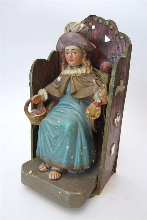 A painted, plaster figurine of a light-skinned child in a blue robe, tan cloak with a white collar, and brown hat is carved on a wooden throne. The child wears a stoic expression with large, placid brown eyes. He holds a basket of fruit.