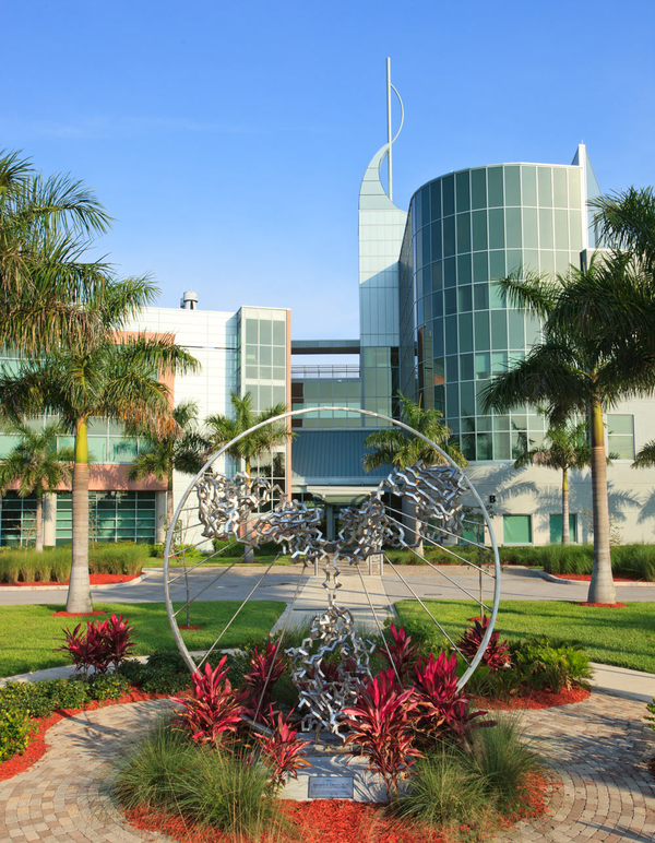 Lines of jagged twisted metal form a sculpture of abstract wings on a bodylike shape. Thin metal spokes emanate from the figure to connect it a large, encircling metal ring. A small garden surrounds the work and a business complex stands in the back. 