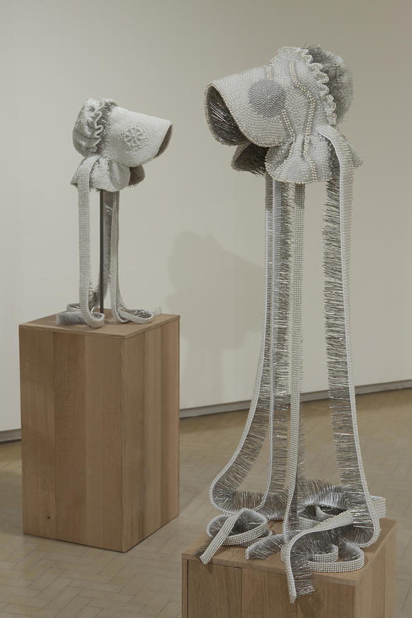 A set of white bonnet sculptures sit on wood platforms. Their expansive brims and long straps are decorated with thousands of corsage pins whose sharp ends point inward.