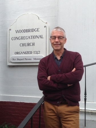 A smiling, light-skinned man in a red sweater and glasses stands against a railing in front of a sign that reads, "Woodbridge Congregational Church Organized 1742."