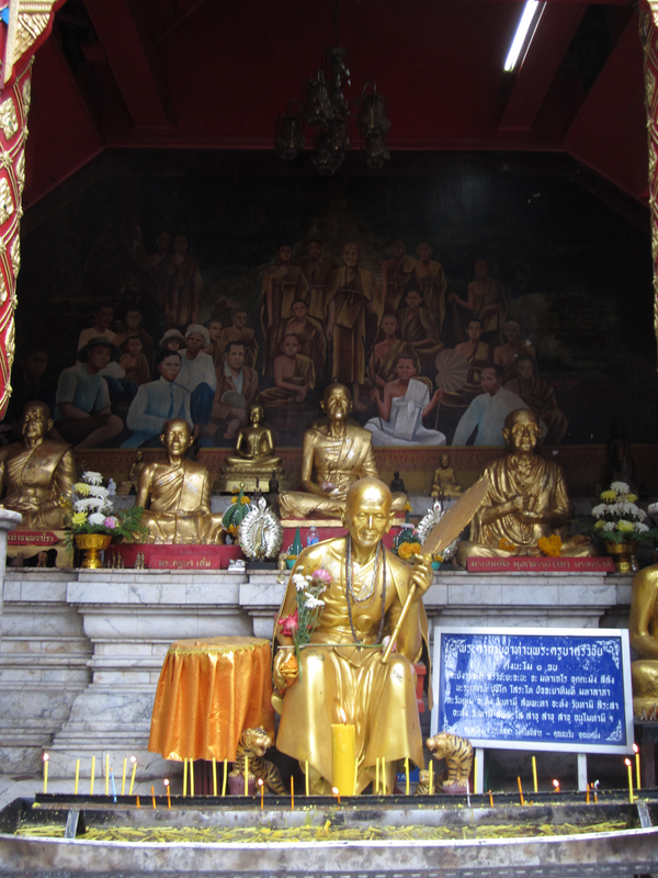 At the center of a shrine sits a golden sculpture of a monk. He wears flowing robes and holds a fan. Necklaces have been draped over his neck and flowers left on his lap as offerings. Sculptures of cross-legged gold monks are sit behind him.
