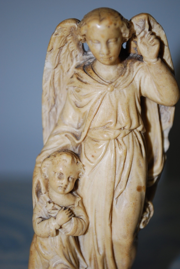A small, unpainted figure of an angel pulls a child close under the crook of its right arm. The angel has large wings, and both figures wear flowing robes. The pair also share a waxy and off-white appearance.