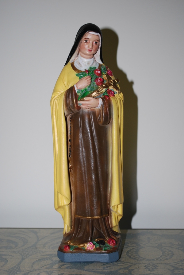 A light-skinned woman wearing a brown habit and black veil holds red roses and a gold crucifix in this plaster figurine. A yellow cape hangs from her shoulders and more roses lie at her feet.