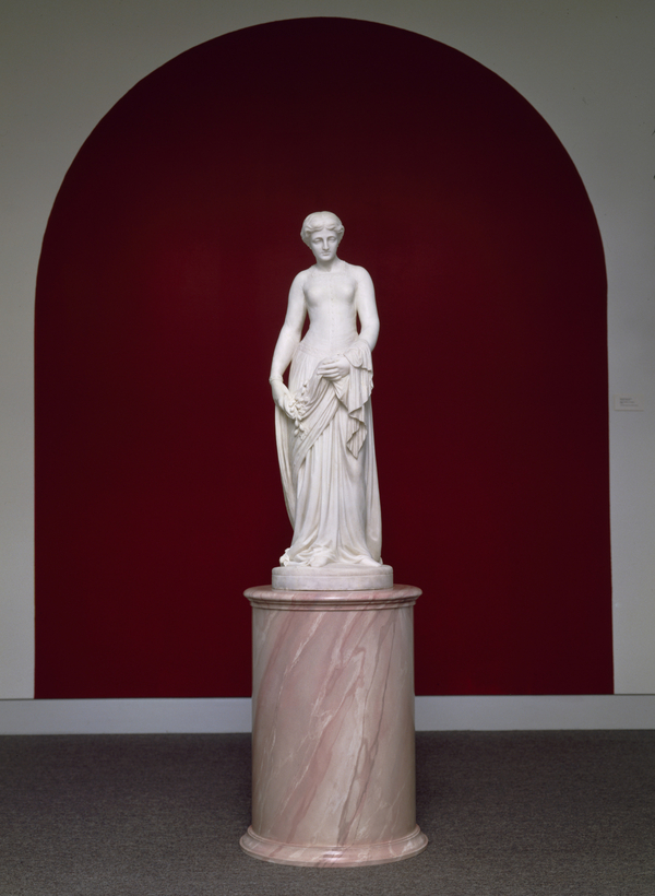 A pale white statue of a thin woman draped in a dress stands on a pink marble pedestal. The work is located against a red wall in a spare room.