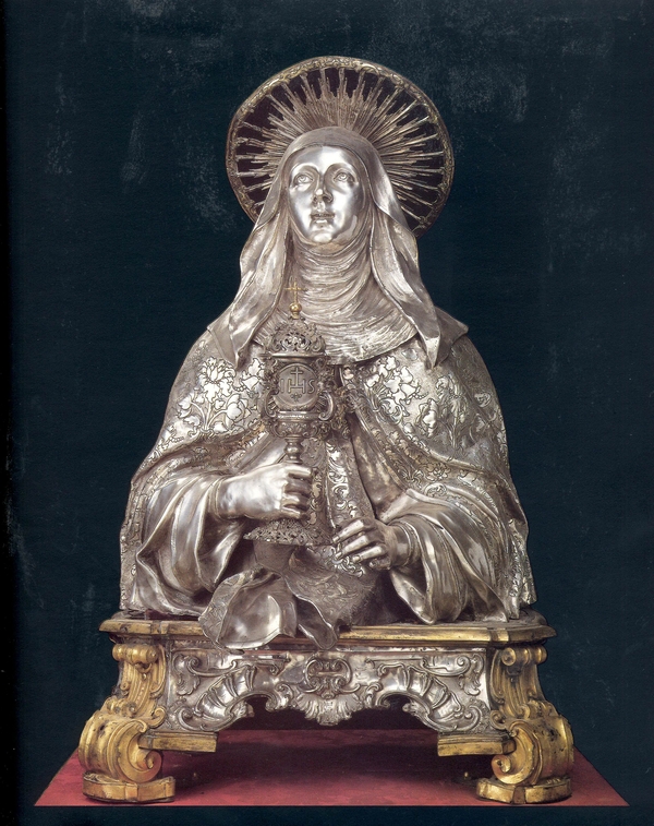 A silver reliquary depicts a bust of Saint Clare holding a monstrance. Her cloak is engraved with flowers and she has a wimple and round halo. She gazes up with bright open eyes.