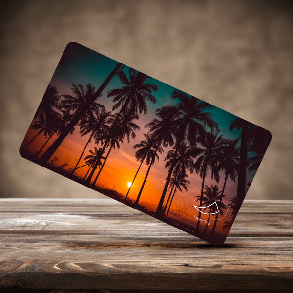 Pictured on a wooden surface, this small rectangular card features a vibrant sunset with a crowd of palm trees in silhouette. In the bottom right corner is a horn-like white outline of a tooth eclipsing a circle, with the words "Tabua Club" written below.