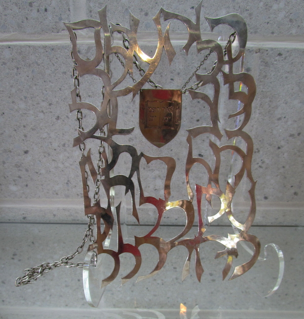 A silver metal sheet formed of swirling filigree frames a smaller metal shield. It hangs on a chair and is engraved with an image of an open book with Hebrew text. The whole piece hangs on more chains like a piece of jewelry.