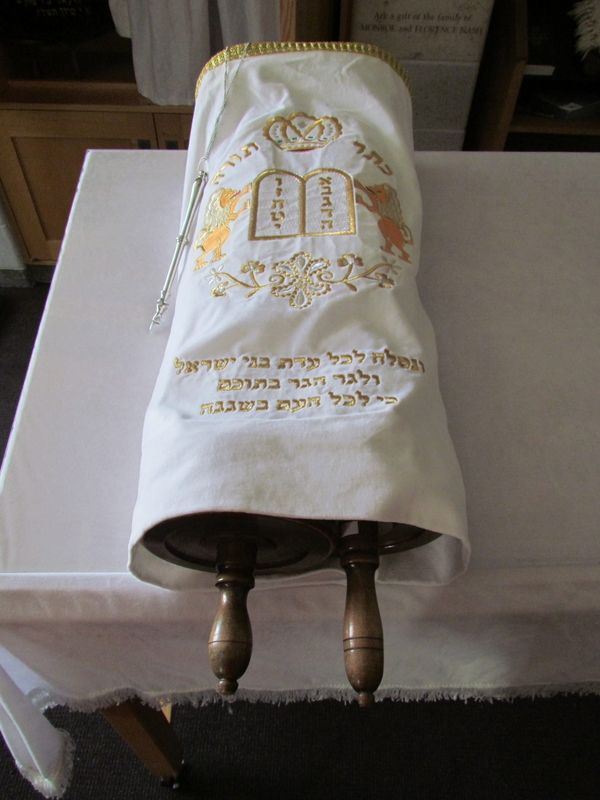 A rolled Torah scroll with two handles is held in a white cloth covering. The cloth is embroidered with gold Hebrew text, a crown, and two lions holding tablets. A silver pointer rests on the same table as the Torah.