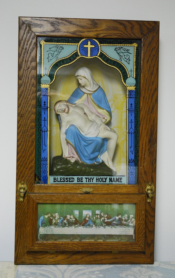 A wooden cabinet with a glass window holds a plaster deposition scene inside. Jesus's white body is cradled by Mary in a blue cloak. A blue decorative border on the glass reads, "Blessed Be Thy Holy Name." A door below has an image of the Last Supper.