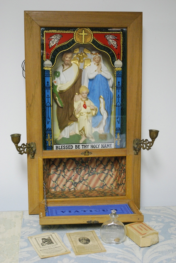 The top half of a cabinet holds a plaster tableau of a light-skinned holy family behind glass. The lower half of the cabinet folds open to reveal a small niche. Prayer cards, a glass bottle, and absorbent cotton are arrayed in front of the open door.