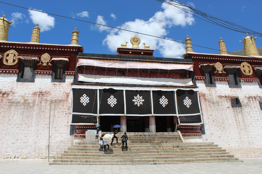 Tibetan monastery of white brick against a blue sky. A small group of women crowd on the stairs leading to the entrance.